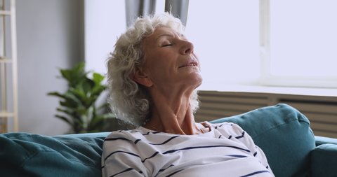 Head shot peaceful mature retired woman breathing fresh air full breasts, relaxing on comfortable sofa alone at home. Mindful happy middle aged tranquil grandmother enjoying break pause leisure time.