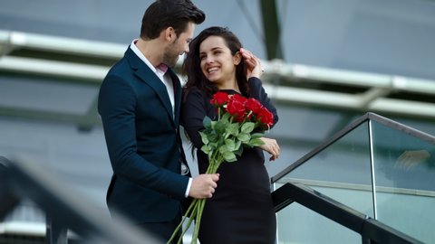 Happy girl waiting for guy in black dress outdoors. Romantic couple hugging with red roses at street. Young couple smiling in stylish clothing together outdoors.