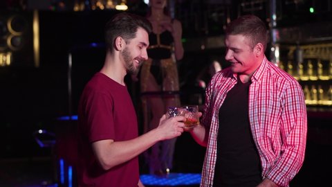 Two young Caucasian men clinking glasses and drinking alcohol in night club. Male friends resting with PJ dancing at the background. Lifestyle, leisure, joy.