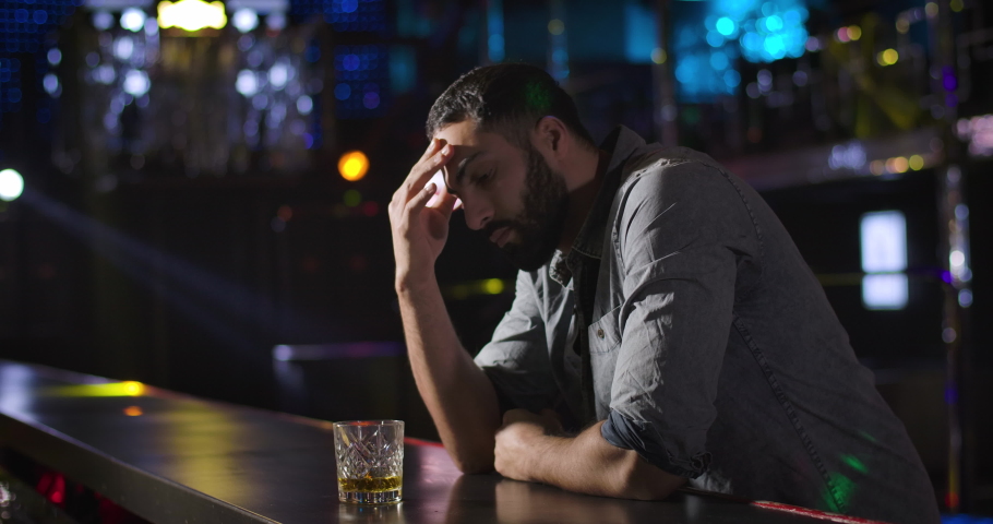 Portrait of depressed lonely Middle Eastern man standing next to bar counter and drinking from glass in night club. One handsome guy spending evening alone. Cinema 4k ProRes HQ. Royalty-Free Stock Footage #1046621716