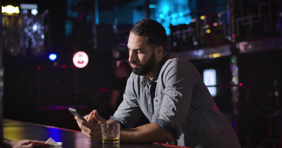 Young Middle Eastern man using smartphone and drinking alcohol next to bar counter in night club. Bored handsome guy resting alone. Cinema 4k ProRes HQ. Royalty-Free Stock Footage #1046621722