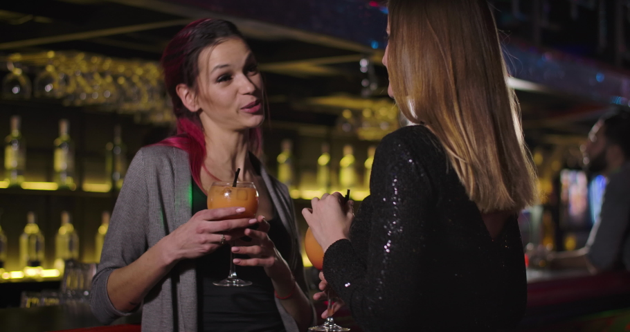 Close-up of two cheerful young Caucasian women dancing with cocktails and talking in night club. Happy girls hanging out and drinking alcohol. Leisure, entertainment, lifestyle. Cinema 4k proRes HQ.