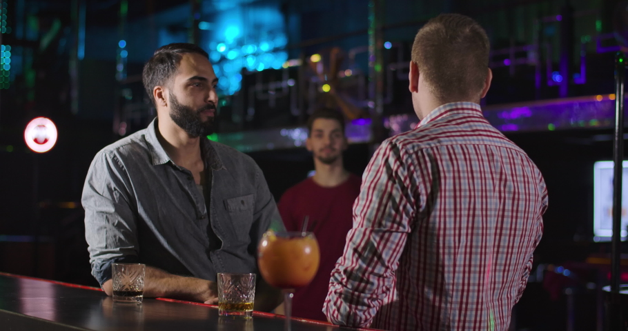 Young smiling Caucasian man coming to fellows in night club and greeting them. Three male friends standing in disco at bar counter and talking. Lifestyle, entertainment, resting. Cinema 4k ProRes HQ.