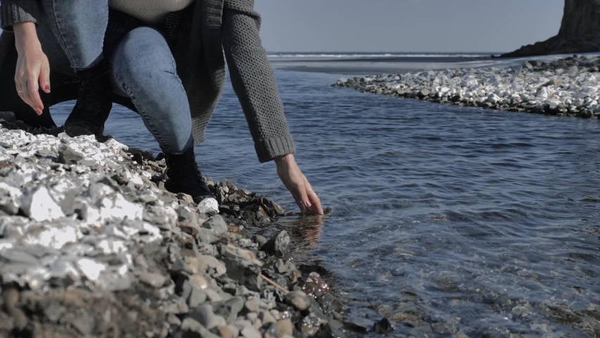 Young girl moisturizes her hand, dipping in clear water on a rocky shore, sunny day
