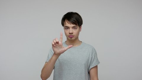 Annoyed dissatisfied brunette guy in T-shirt showing loser gesture, making L sign with fingers, pointing to camera meaning you lost, blaming for failure. indoor studio shot isolated on gray background