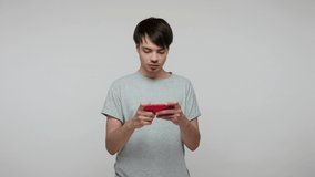 Entertainment, leisure and technology. Positive guy in t-shirt playing video game with interest, using mobile phone, enjoying application, online gaming. indoor studio shot isolated on gray background