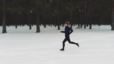 Running Man In Sportswear Workout Before Triathlon. Runner Man Running Winter Forest Snow Trail. Winter Sports And Recreation Concept. The camera flies to the side of the runner in the winter forest