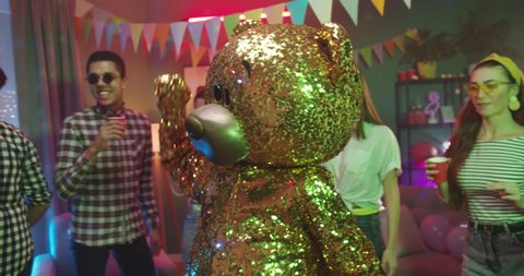 Big golden grown doll of teddy bear dancing funny in the middle of the friends in the decorated room at home party. Indoors.