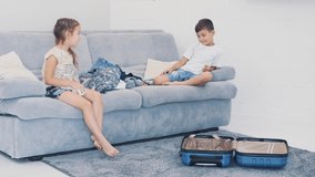 Little children are throwing clothes into suitcase at home. They are sitting at the blue sofa. Closeup 4K.