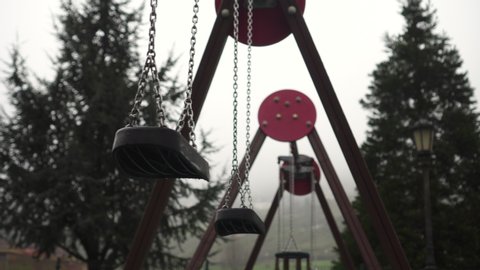 Seats of a swing with chains swing in foggy cloudy weather in a park with coniferous trees. Dramatic look. Coronavirus quarantine concept