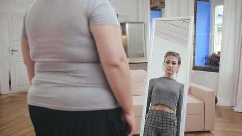Happy fat woman in sportswear looking at fit reflection. Female confident person concept. Happy plus size fat girl looks at herself in mirror and seeing slim reflection. Happy body-positive concept.