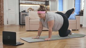 Fitness online. Overweight young woman doing exercises for weight loss at home in the morning, using laptop. Obese woman exercising yoga alone on the floor in house watching tutorial video