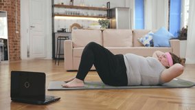 Home fitness concept. Overweight woman doing strength training abs bodyweight floor exercises for weight loss watching dvd workout or web videos on laptop in living room