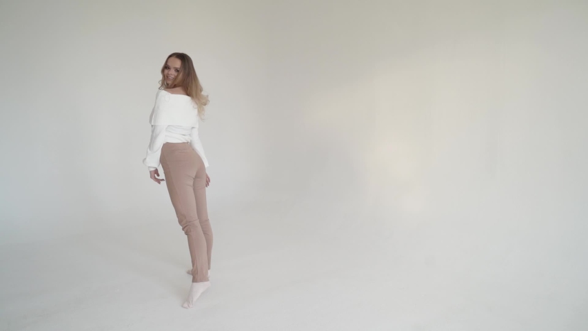 beautiful young blonde girl in a white blouse and light trousers makes a turn with her body on a white cyclorama, brings a finger to her lips and makes a wave with her hair