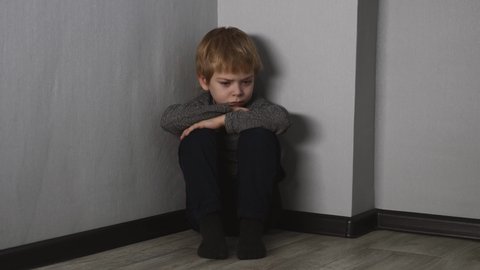 A scared, lonely blond boy sits on the floor in the corner of the room.