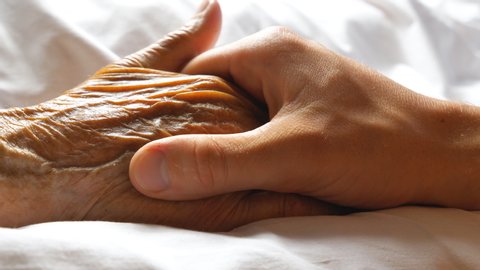 Worried man taking and gently stroking hand of his sick mother showing care or love. Son comforting wrinkled arm of elderly mom lying at bed. Guy giving support to his old parent. Side view Close up