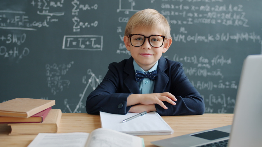 Portrait of little genius adorable boy in classroom sitting at desk wearing glasses smiling looking at camera, a chalkboard with formulae is in background.