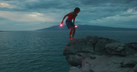 Extreme sports cliff diver doing a backflip off of a sea cliff with burning red flares, epic video of guy being awesome