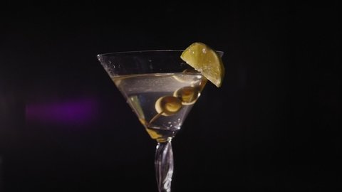 Martini is poured into a glass with olives and lime which stands on a rotating stand on a black background. Slow motion. Artificial lighting.