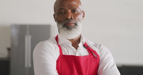Portrait close up of a senior African American man with a white beard, wearing a red apron, standing in cookery class with arms crossed, looking to camera and smiling, in slow motion