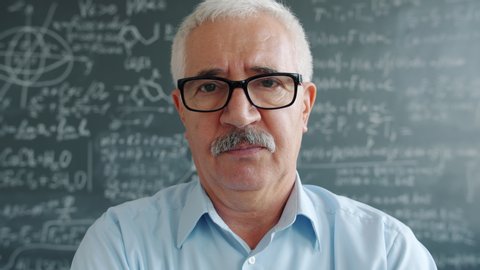 Portrait of smart man researcher standing indoors in class looking at camera with serious face, chalkboard with formulas is visible in background. People and science concept,