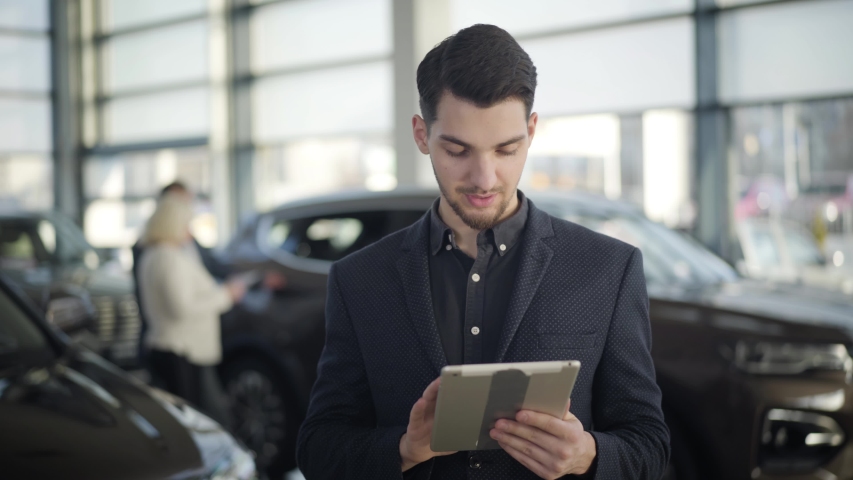 Young handsome brown-eyed Caucasian man standing in car dealership with tablet, looking at camera and smiling. Confident brunette dealer posing at workplace in showroom. Job, occupation, lifestyle. Royalty-Free Stock Footage #1046723491