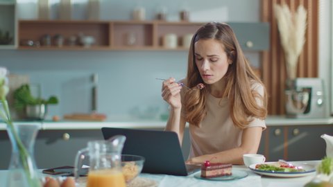 Businesswoman working on laptop computer at home office. Portrait of thoughtful girl eating cake on domestic kitchen. Pretty business woman using laptop on kitchen in slow motion