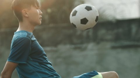 Young Korean guy with blue jersey practicing soccer skills and tricks with the ball at sunset in an outdoor urban spot with grungy walls