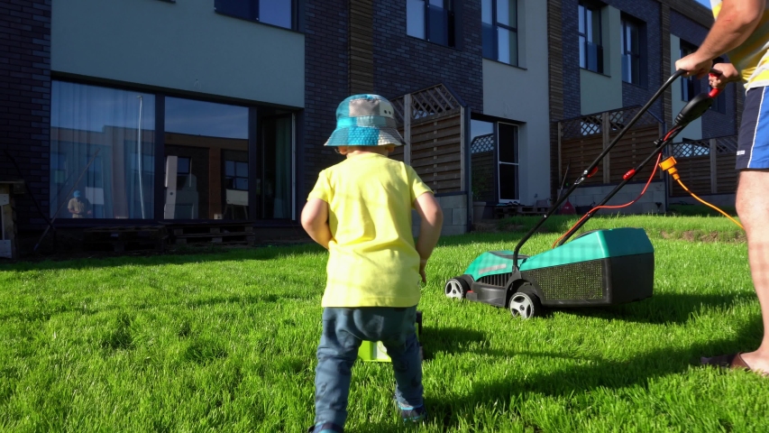 Father and son mow lawn in private yard. Funny scene of man and boy working with lawn mowers. Gimbal movement shot. Royalty-Free Stock Footage #1046739289