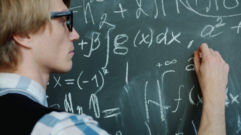 Young mathematician is busy writing formulas on chalkboard working with serious face wearing glasses and casual clothing. People and occupation concept.