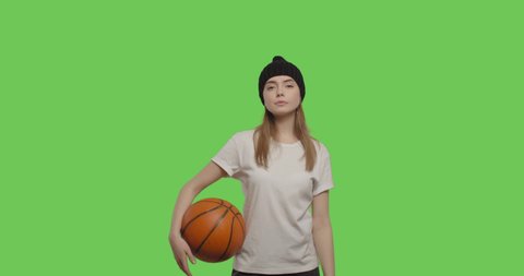 Girl wearing white t-shirt and black beanie posing over green screen background. Young woman holding basketball ball on Chroma Key. 4k raw video footage slow motion 60 fps