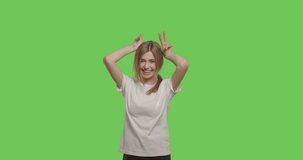 Smiling young crazzy woman having fun and making horns by her hands, looking at camera over green screen background. Girl showing victory sign on chroma key. 4k raw video footage slow motion 60 fps