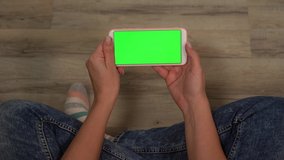 Closeup point of view of woman sitting on wooden floor and looking at empty green screen of modern smart phone.