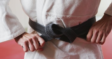 Front view mid section of a Caucasian male judoka standing on gym mats, tying up the black belt of white judogi in slow motion