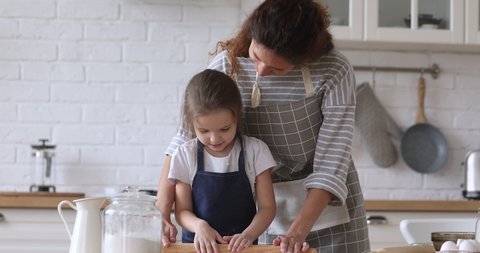 Happy young mommy helping little preschool daughter using rolling pin, preparing dough for homemade pastry together in modern kitchen. Smiling small kid girl enjoying cooking baking with mom.