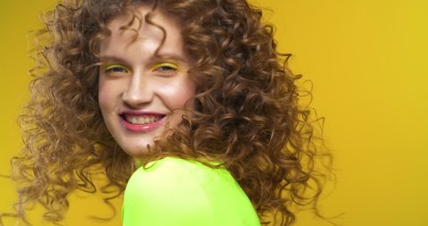Young woman with natural Beauty and Colorful Makeup posing on Camera at Studio. Looking stylish and wearing trendy Clothes. Having fun and turning while her Curly Hair waving. Glamorous lipstick.