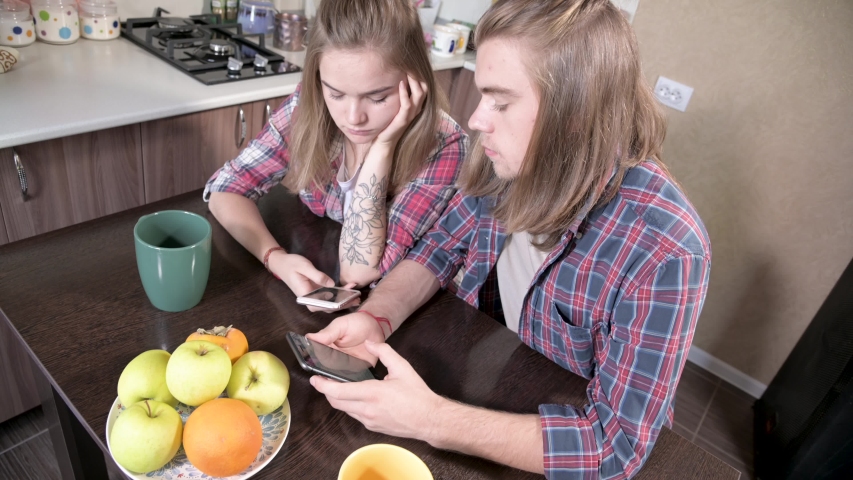 A young pair of mellenials with long hair are sitting at the kitchen table with phones in their hands. Surfing and shopping on the Internet from mobile devices. Concept of modern young family