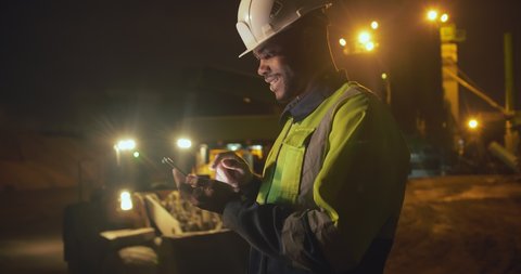 Afro-american smiling supervisor using mobile phone while standing outside concrete manufacturing carrying out inspecting of night shift workers. Industrial worker with cellphone at night at work