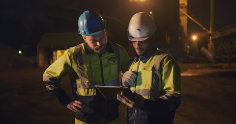 Multiethnic builders in hardhats and safety uniform with tablet computer outdoors on construction site at night shift. Stone quarry managers working night shift