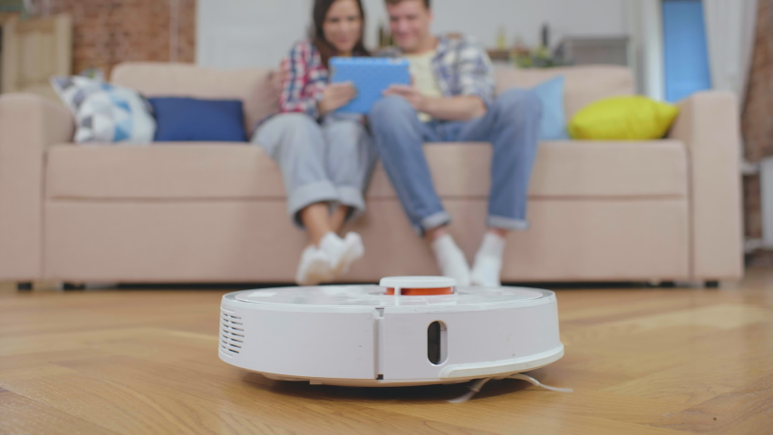 Young happy husband and wife relaxing in living room and shopping online on tablet computer while modern vacuum robot cleaning floor. Automatic housecleaning and modern technology concept.