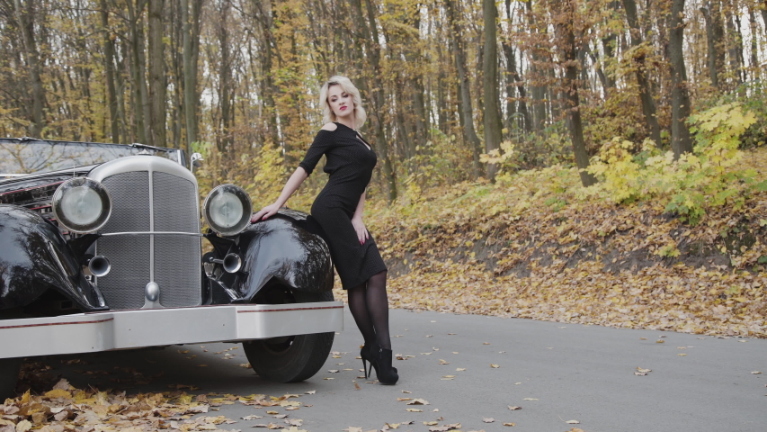 Portrait of passionate blonde in black dress stands in sexy pose at retro cabriolet in autumn forest. 4K