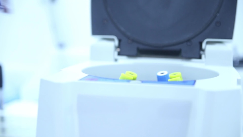 lab technician wearing a white lab coat and blue latex gloves adding blood sample test tube to alab testing device 