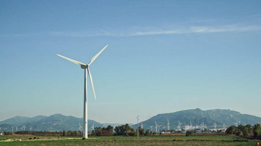 Locked down shot row left to right of windmills on green meadow, under bright blue sky on background of mountains in Spain. Working electric modern new windmills in Tarifa