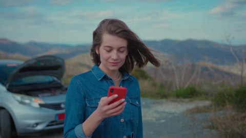 Young upset woman standing at broken car searching for repair service on smartphone worrying in stress. Car accident. Car trouble.