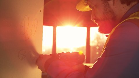 SLOW MOTION, CLOSE UP, LENS FLARE, DOF: Bearded construction site worker working late fastens a bolt inside a wall panel. Golden sunbeams shine on worker mounting a panel of drywall with a power drill
