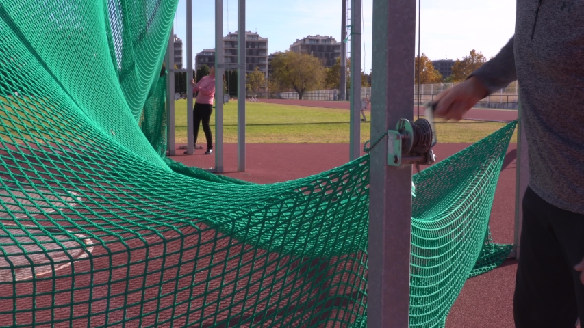 Detail shot of a man with a gray pullover climbing the nets of a running track with one of the pulleys, in the background a woman with another of the pulleys, outdoor athletic stadium