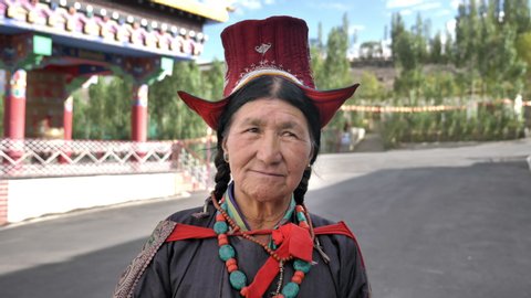 An Old Tibetan woman in traditional costume looking into a camera standing outside Buddhist monastery situated in Himalaya Mountain. A smiling elderly lady wearing ethnic head wear.