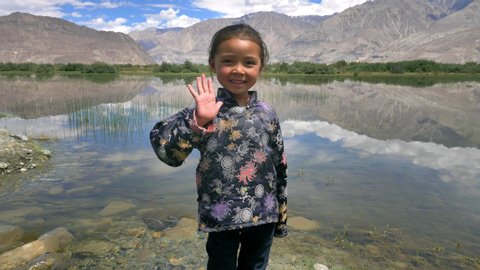 A happy cute girl is smiling and waving looking at the camera standing against mountains and blue skyClouds reflecting in the clear water. An adorable AsianTibetan kid with a smile on her face. 