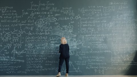 Time-lapse of female scientist blonde in elegant suit writing formulas on chalkboard working alone concentrated on theory. People and education concept.