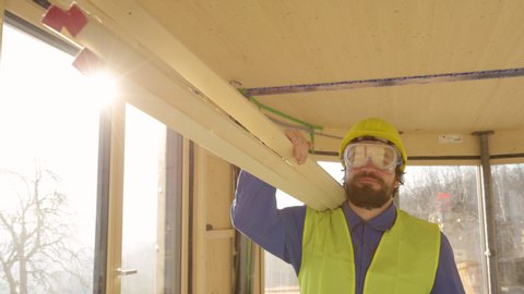 SLOW MOTION, CLOSE UP, LENS FLARE: Smiling contractor in blue workwear carries planks across a glue-laminated house under construction. Young Caucasian worker carrying planks across a sunlit room.